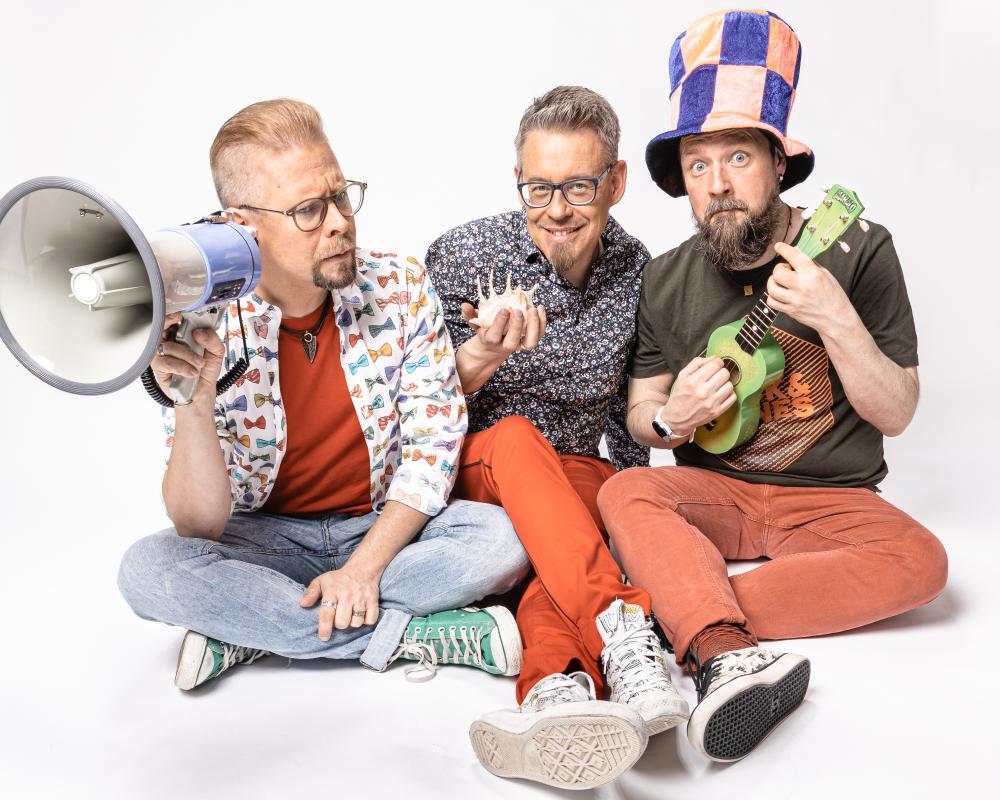 Pekka Laukkarinen trio värikkäissä vaatteissa lattialla otetussa yhteiskuvassa. 