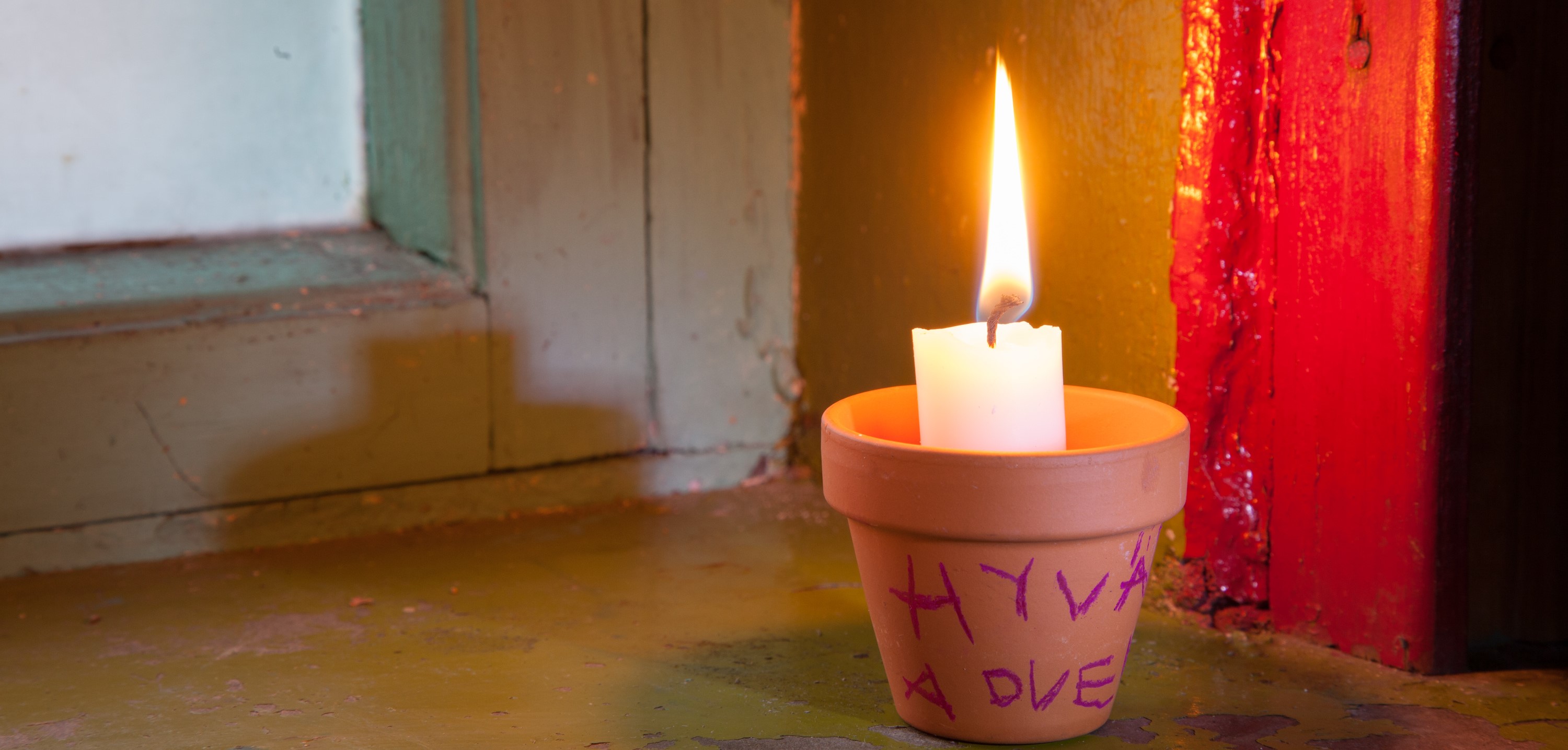 Kukkaruukkuun laitettu palava kynttilä ikkunalla. Ruukussa lapsen kirjoittama teksti hyvää adventtia.
