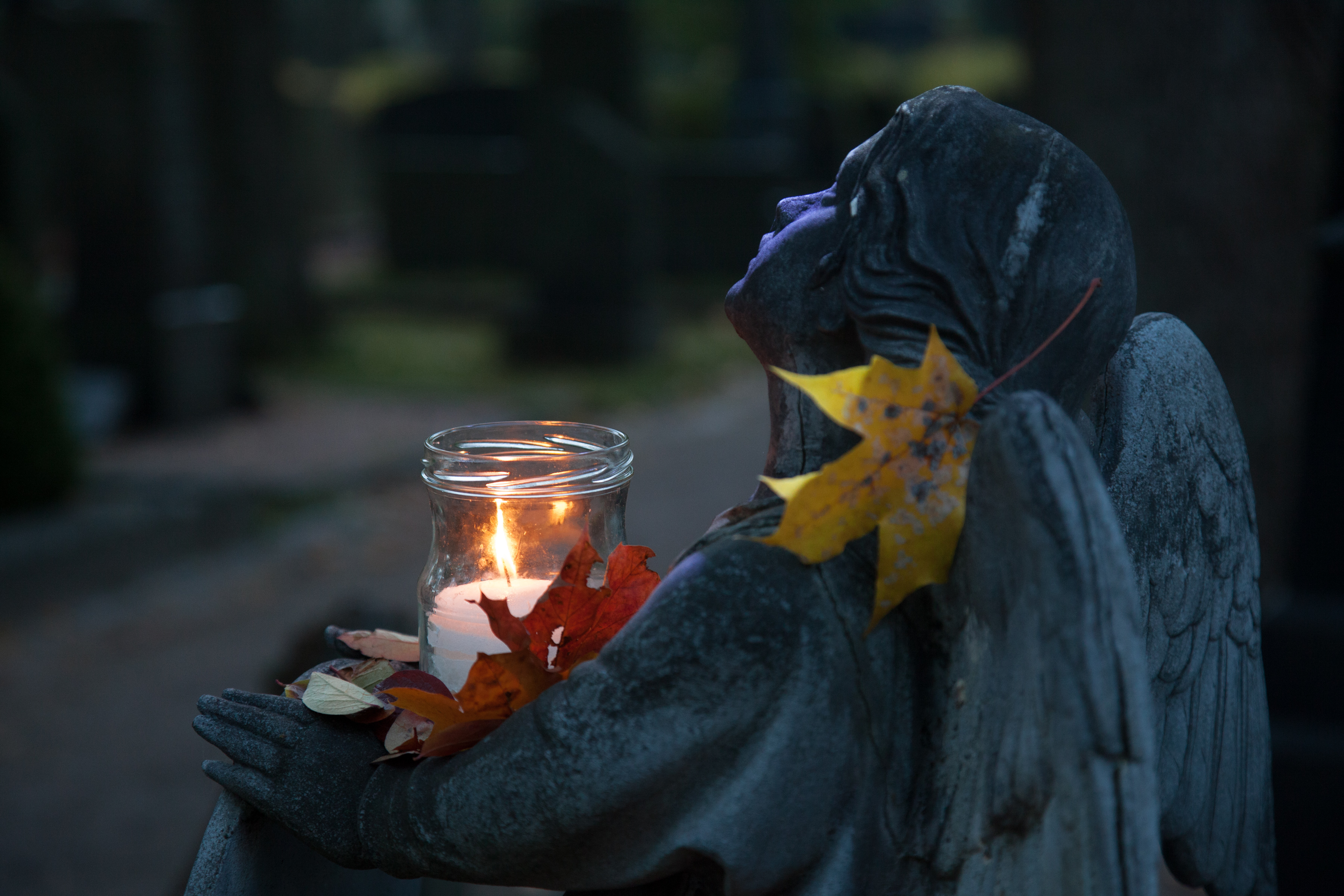 Pyhäinpäivän symbolikuva hämärältä hautausmaalta, jossa edessä enkelipatsas katsoo ylös sylissään kynttiläl...