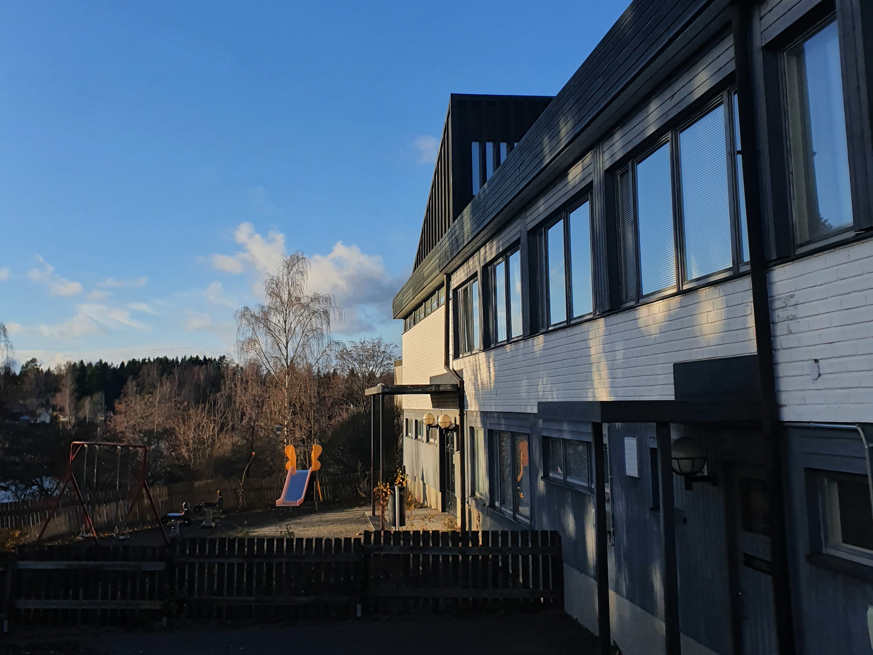 Lempoisten seurakuntatalon vaalea etelän puoleinen pääty. Pihalla on pieni ulkoleikkialue. Taustalla sinine...