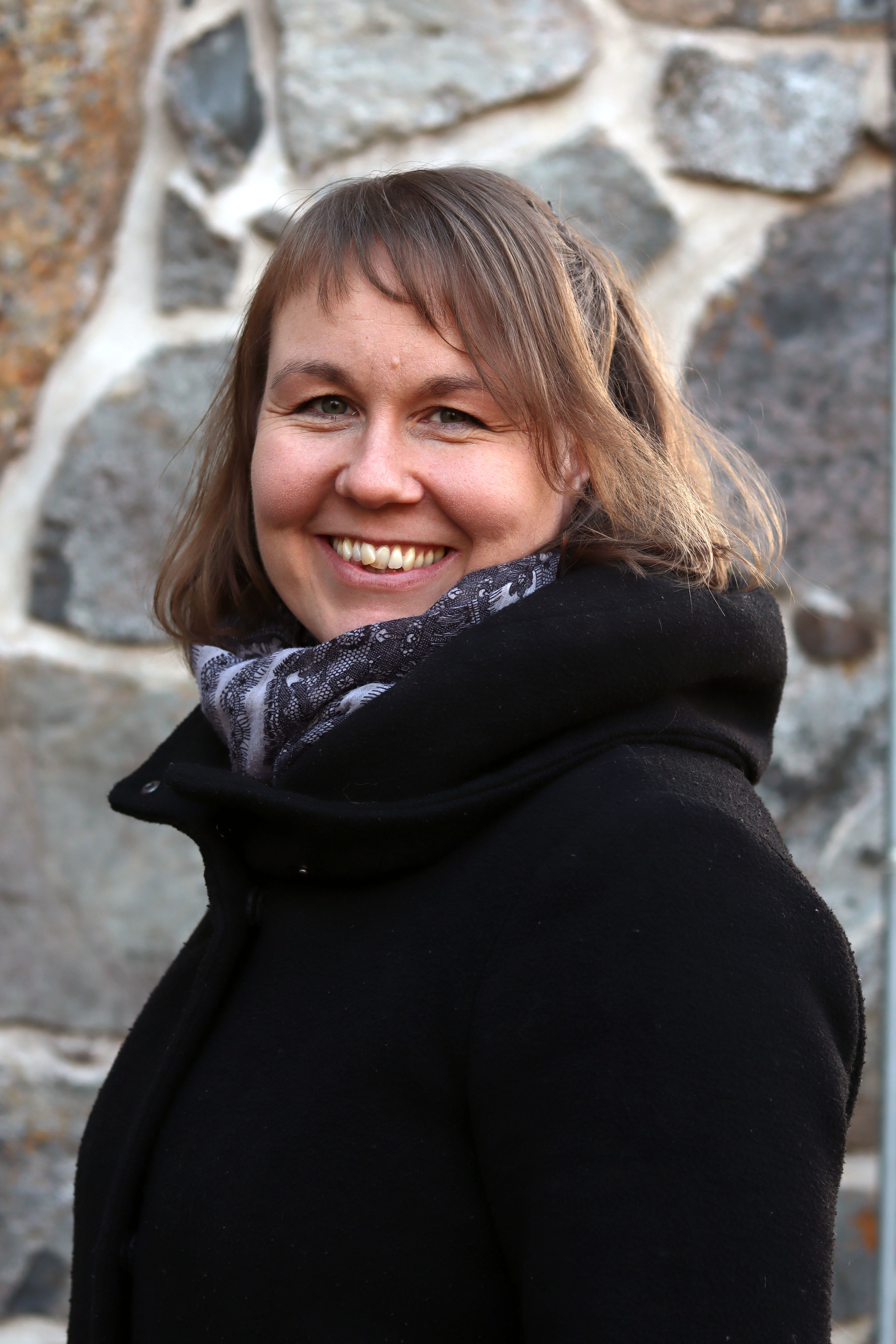 Kanttori Sanna Pelvola hymyilee kirkon kiviseinän edustalla talvivaatteissa.