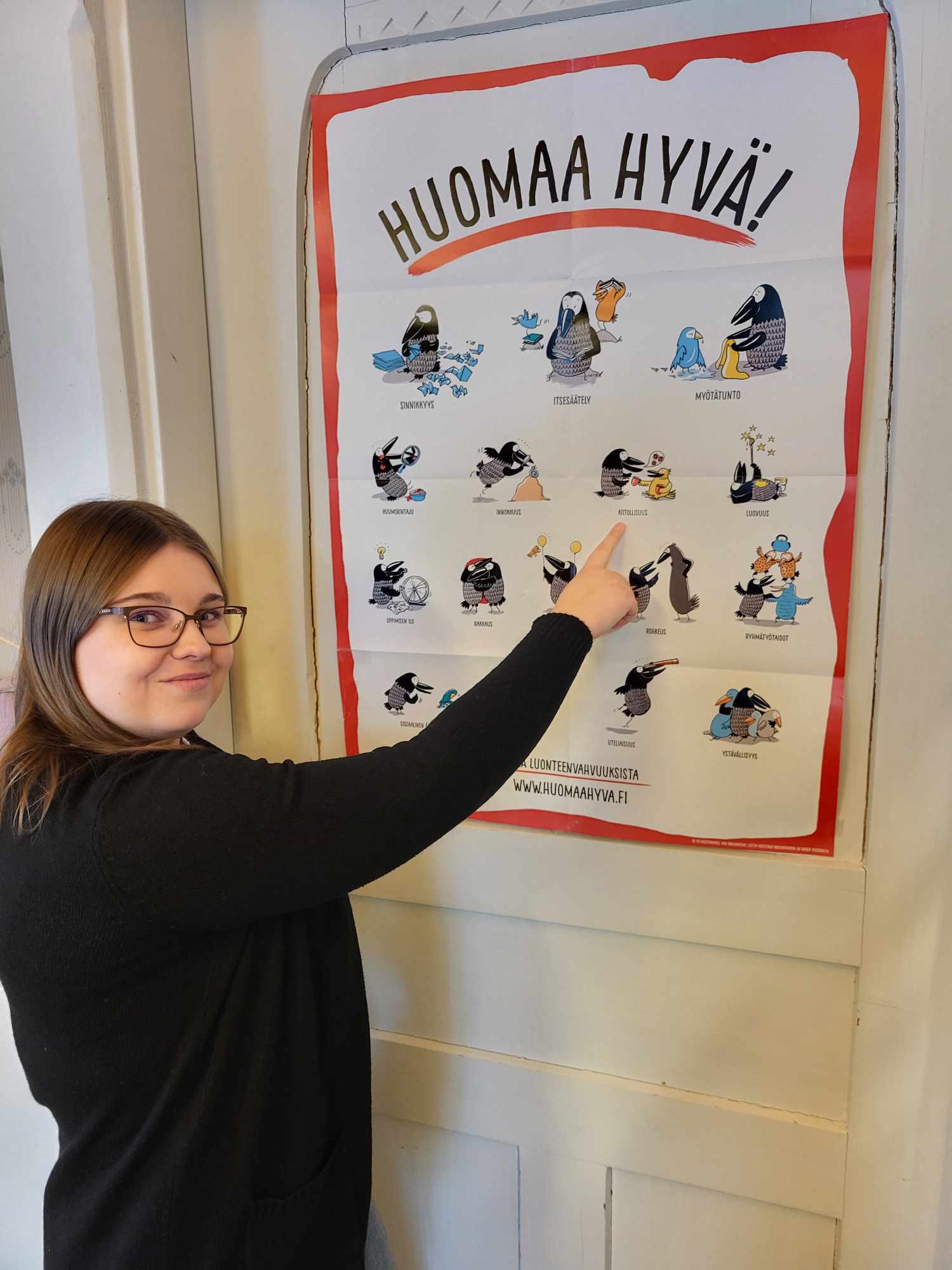 Silmälasipäinen, tummahiuksinen nainen osoittaa Huomaa hyvä -otsikoidun julisteen kuvaa ja tekstiä.