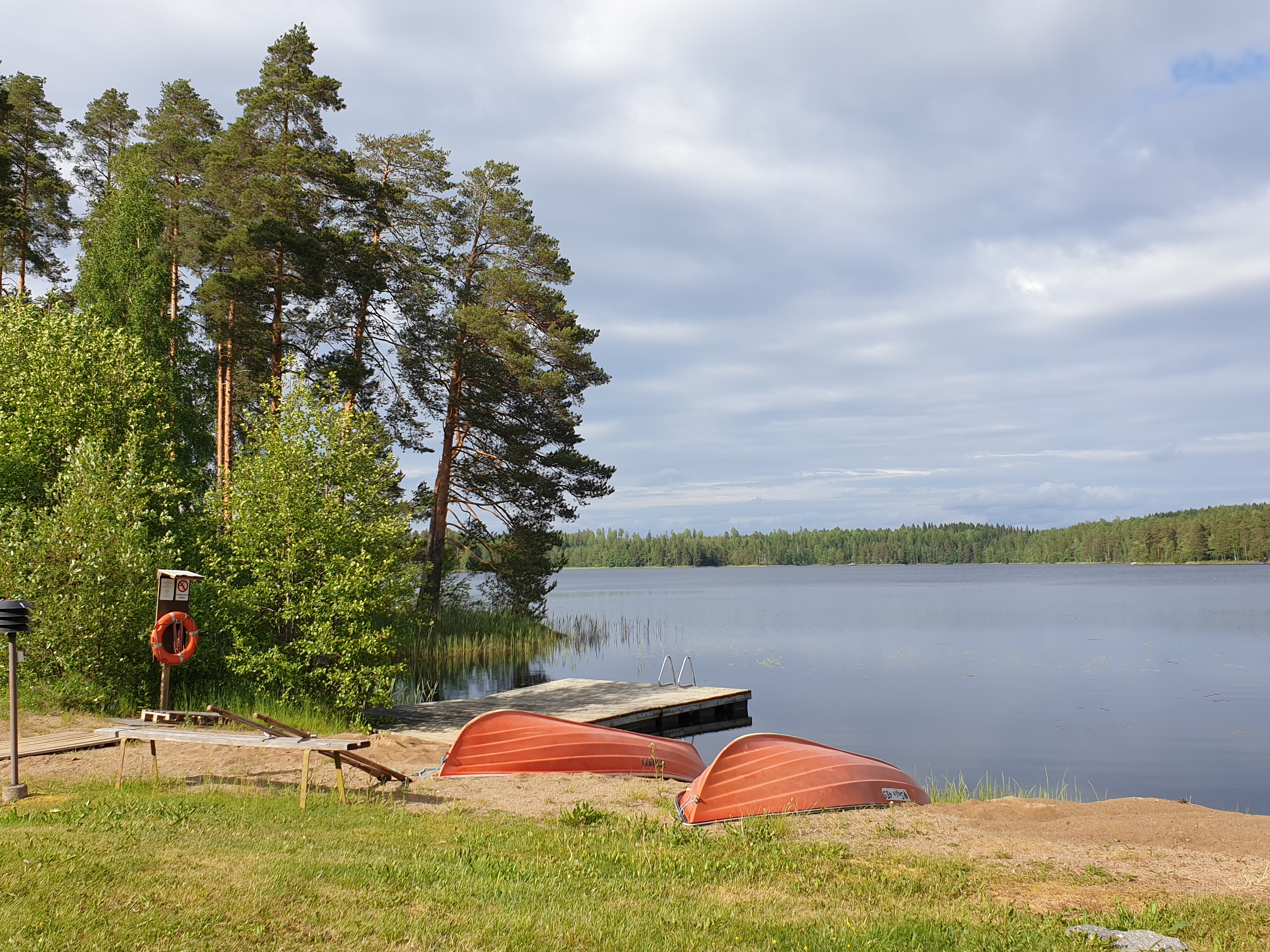 Näkymä Tervajärven leirikeskuksen rannasta: mäntyjä,  järvenlahti, taivasta, hiekkarannalla veneitä, pelast...
