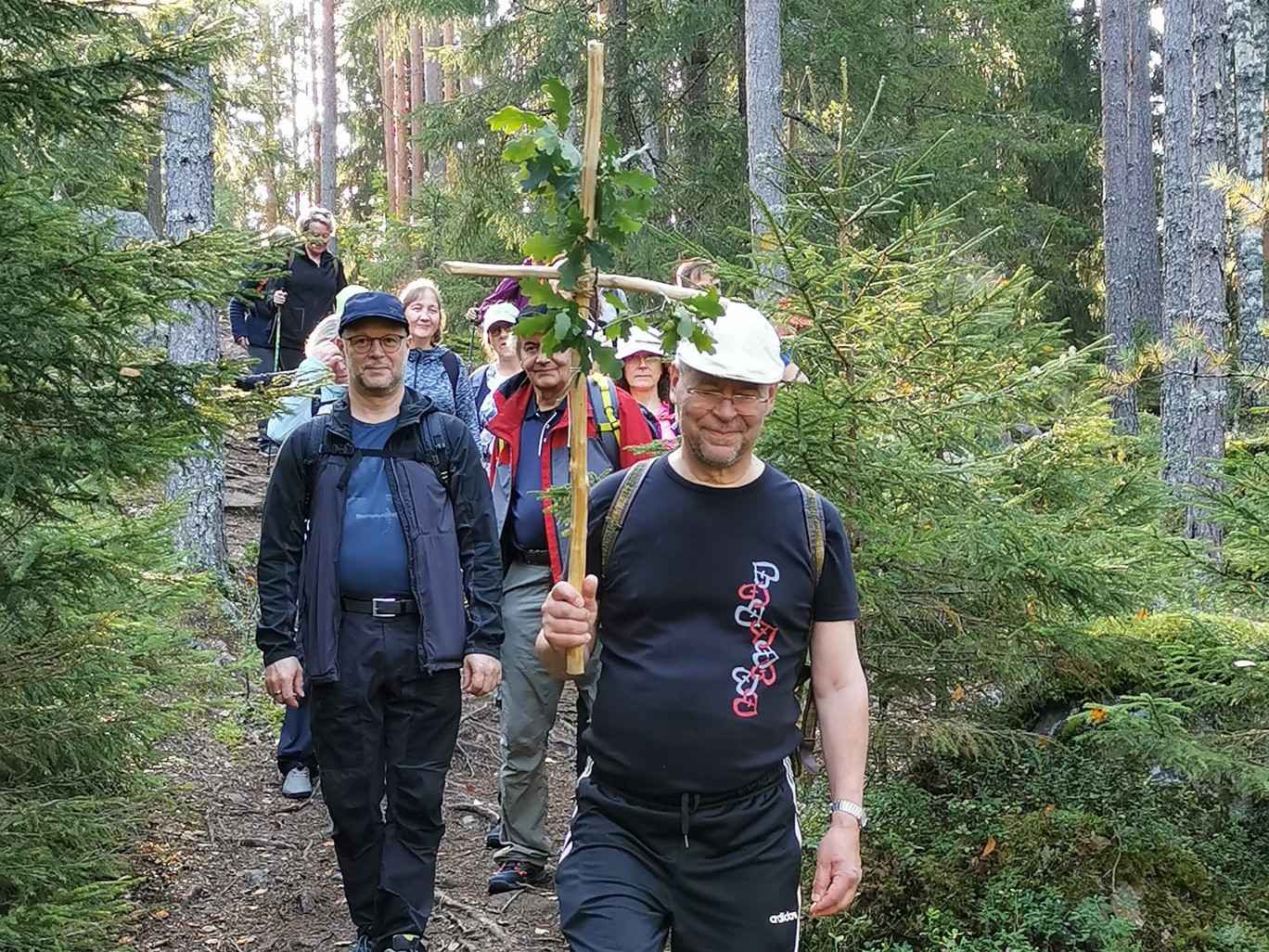 Vaeltajia metsäpolulla. Ensimmäisellä on puinen risti kädessään.