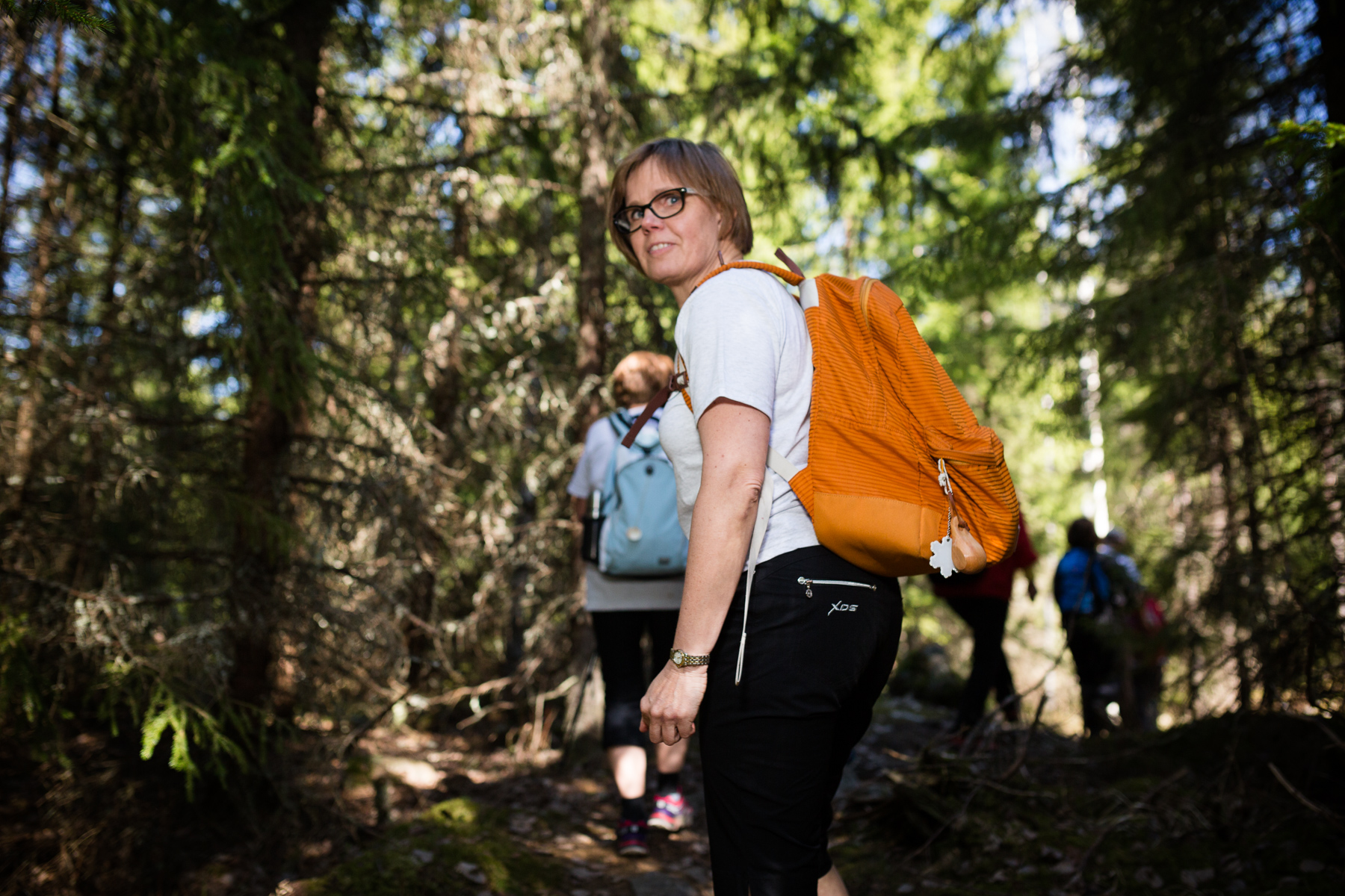 Keltareppuinen naisvaeltaja on lähdössä kohti edessä näkyvää metsäpolkua ja katsoo vielä taakse (kameraan).