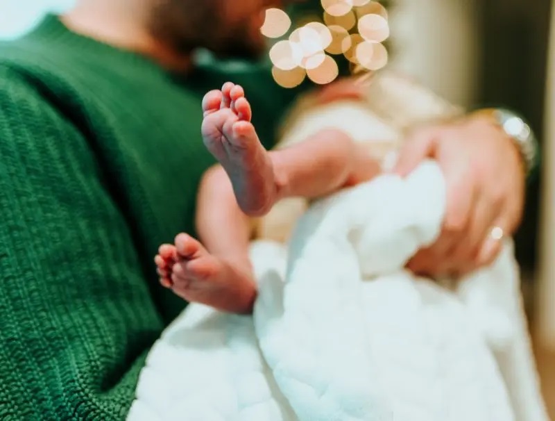 Aikuinen pitelee sylissään lasta. Pienet jalat näkyvät valkoisen, pitkän puvun alla