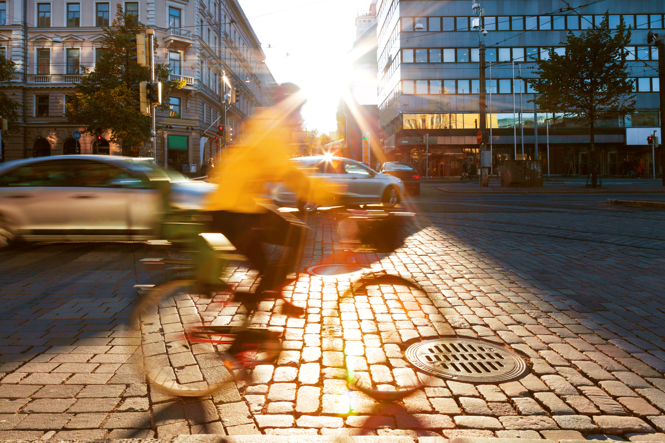 Pyöräilijä ison kaupungin liikenteessä, talojen välistä virtaa auringonvaloa.