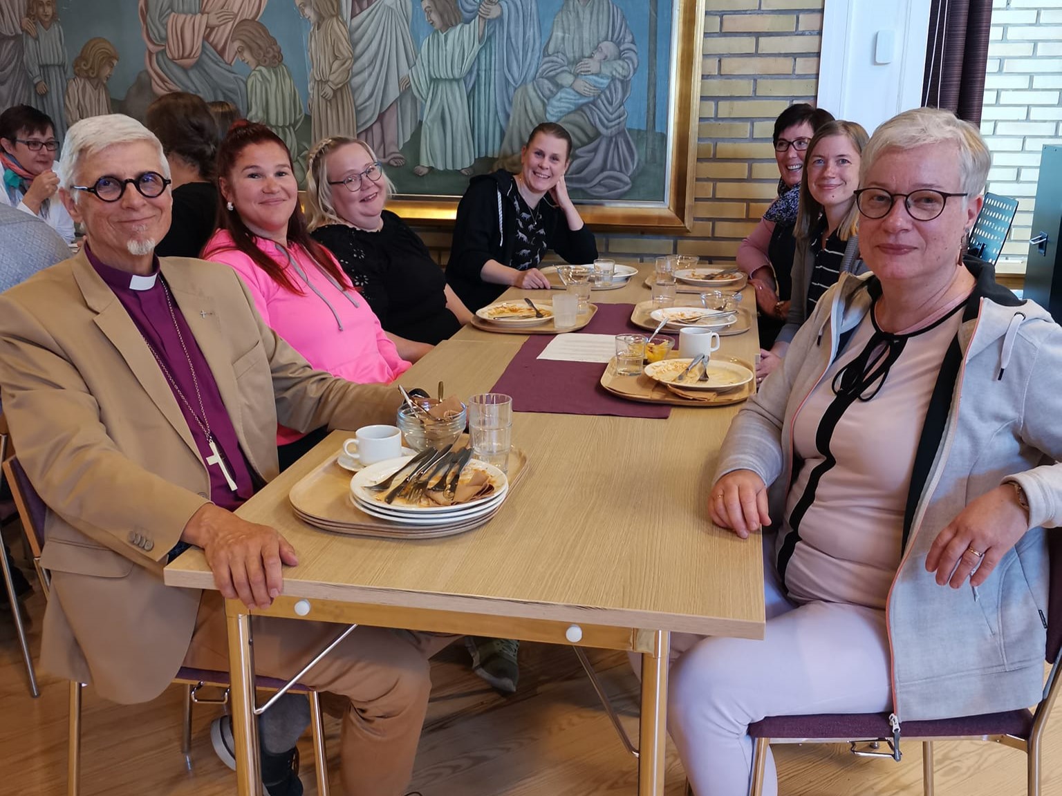 Lounaspöydän ääressä hymyilevä joukko, edessä vasemmalla piispa Matti Repo purppuran värisessä piispanpaida...