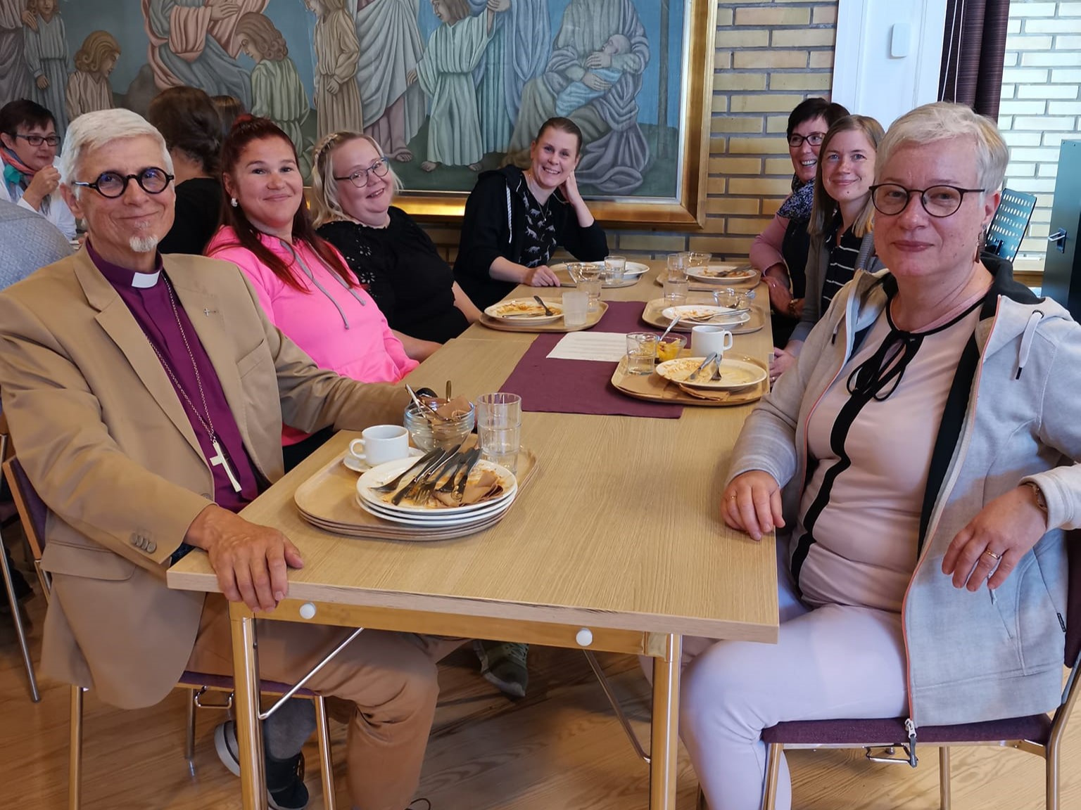 Pitkän lounaspöydän päässä vasemmalla Matti Repo purppuran värisessä piispanpaidassa, pöydässä lisäksi ryhm...