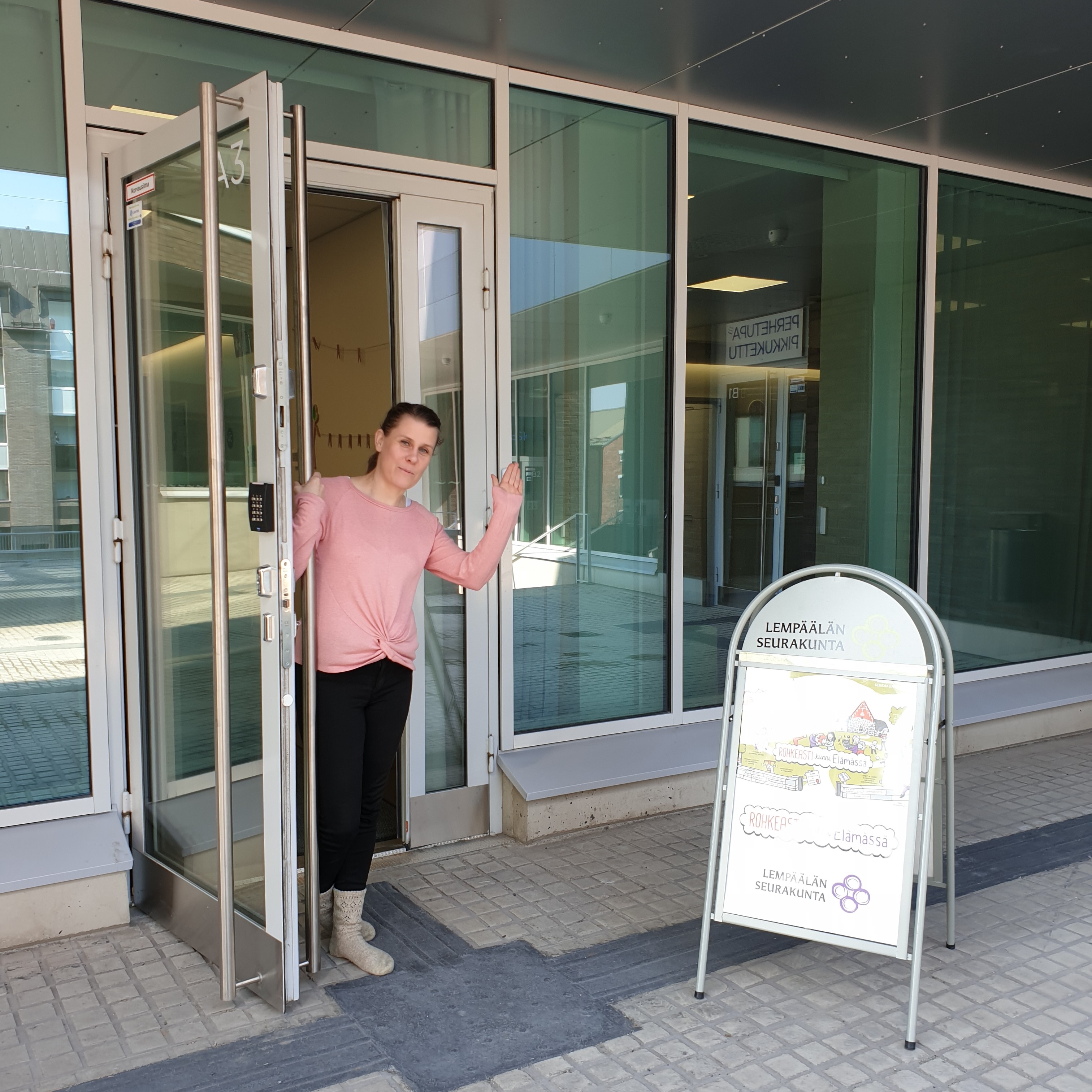 Seurakuntatoimisto Lempäälä-talossa Hymyilevä nainen pitelee seurakuntatoimiston ovea auki ja toivottaa tervetulleeksi.