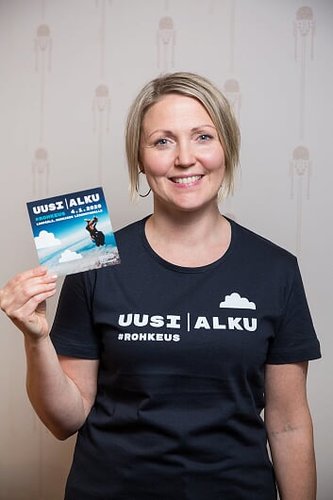 Heli Orre poseeraa Uusi Alku -tapahtuman t-paidassa ja pitelee tapahtuman esitettä kädessään.